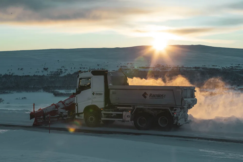 Ramlos-lastebil som kaster snøføyke over vintervei ved solnedgang