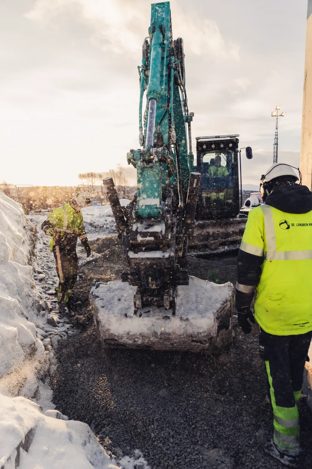To anleggsarbeidere og maskinfører i beltegraver utfører grunnarbeid i grus og snø i lav vintersol