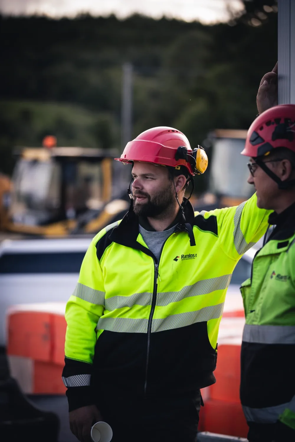 To anleggsarbeidere med røde hjelmer og neongule varseljakker tar pause med kaffe ved anlegget