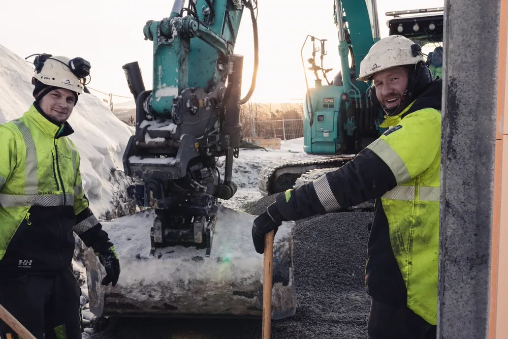 To anleggsarbeidere utfører grunnarbeid foran blågrønn gravemaskin i snødekt vinterlandskap