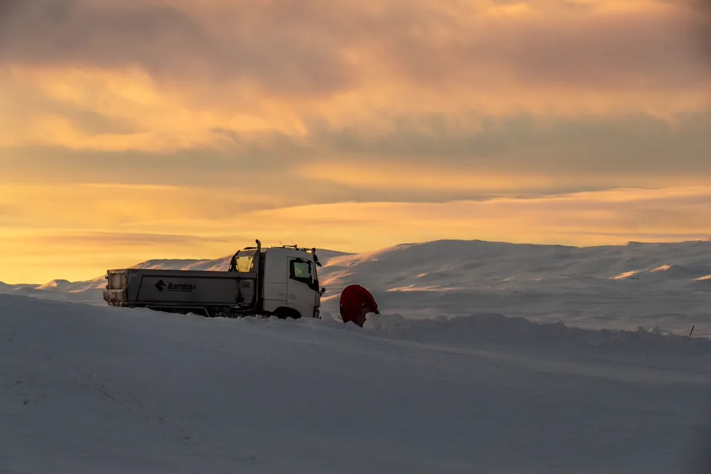 Ramlos-lastebil med rød snøplog står på snødekt fjellvidde under solnedgang