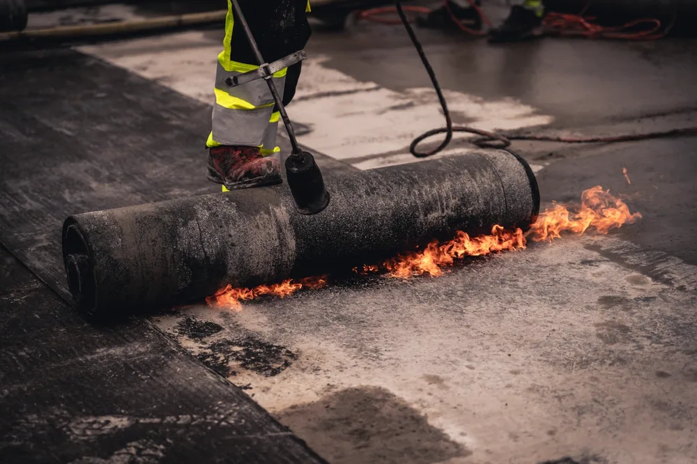 Arbeider sveisser takbelegg med åpen flamme på et flatt betongdekke