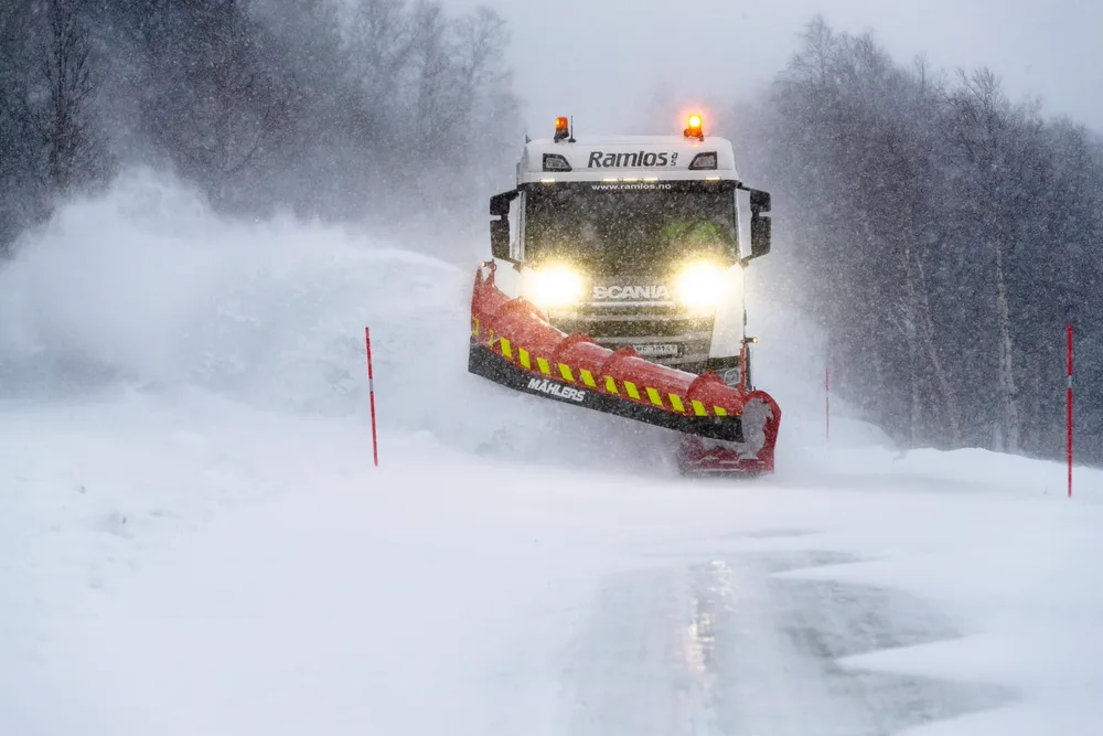 Sjåfør brøyter nedsnødd vei i tett snøvær med hvit Scania-lastebil og rød Mählers-snøplog