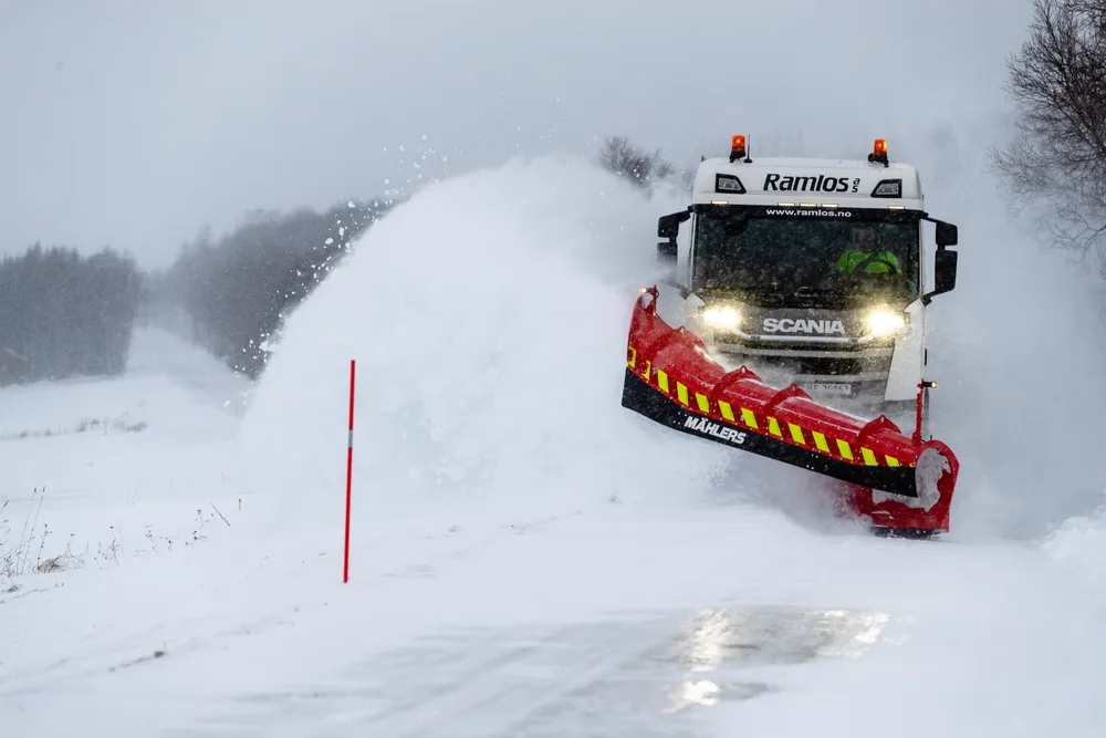Hvit Scania-lastebil med rød og gul Mählers-plog kaster snø til siden i snødekt landskap
