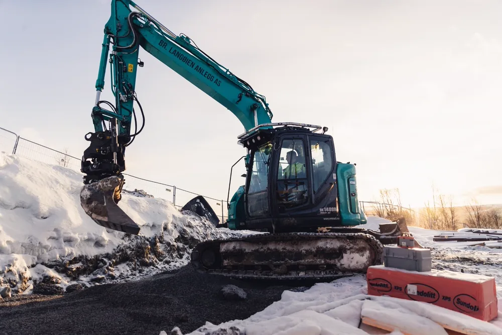 Fører i turkis Kobelco beltegraver fra Br. Langøien Anlegg AS graver på snødekt byggeplass