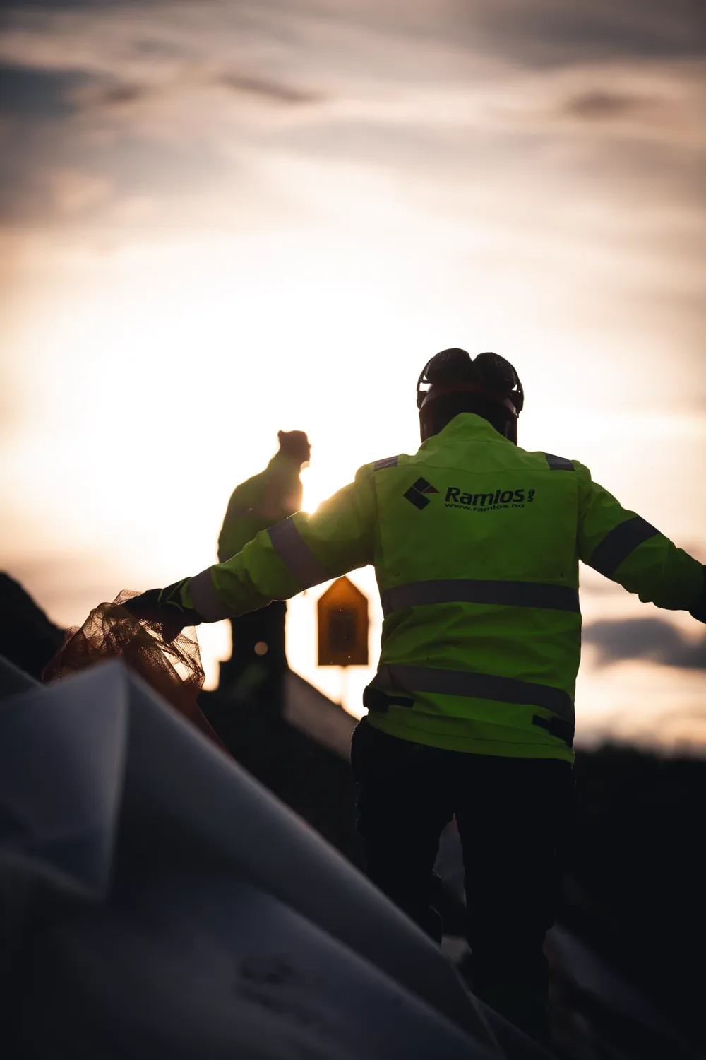 Jernbanearbeider i refleksjakke med Ramlos-logo på ryggen håndterer nettning i motlys ved solnedgang