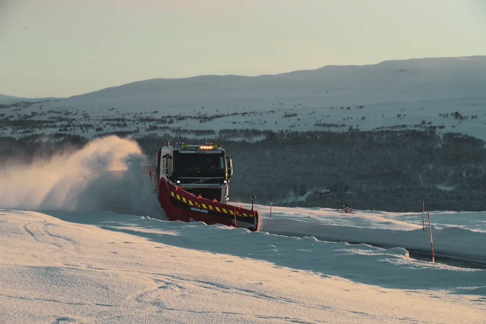 Hvit Volvo-lastebil med rød Mählers-snøplog brøyter fjellvei foran bjørkeskog