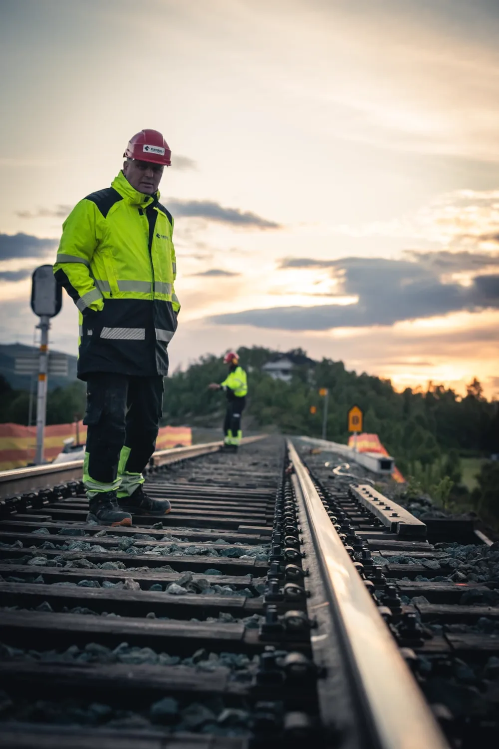 Banearbeider står på jernbaneskinnene med hendene i lommene mens en kollega står lenger bak på traseen