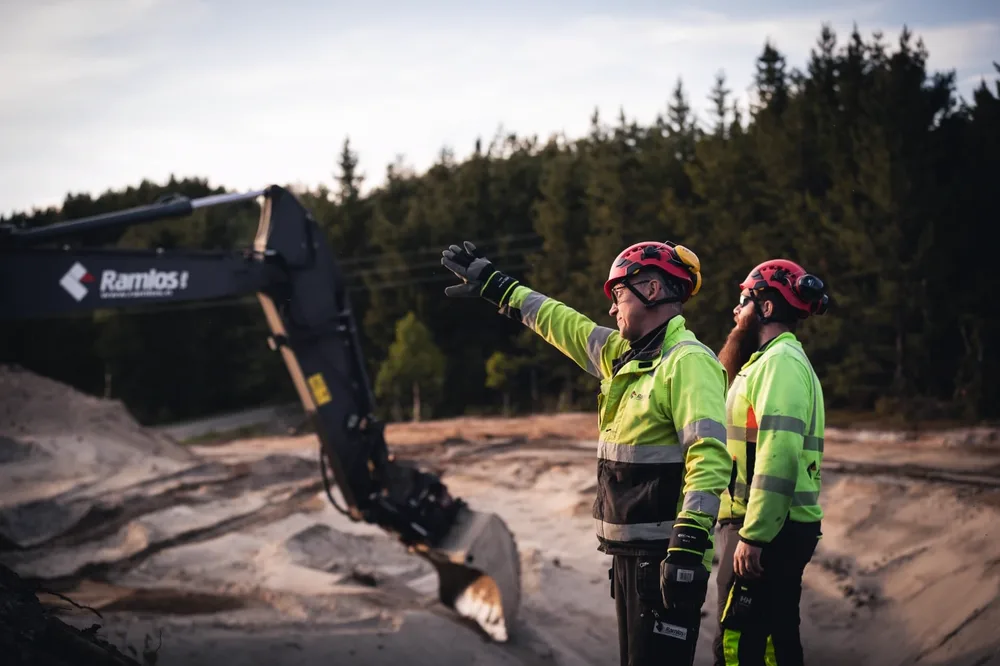 To anleggsarbeidere dirigerer arbeid i sandtak foran gravemaskin med Ramlos-logo på bommen